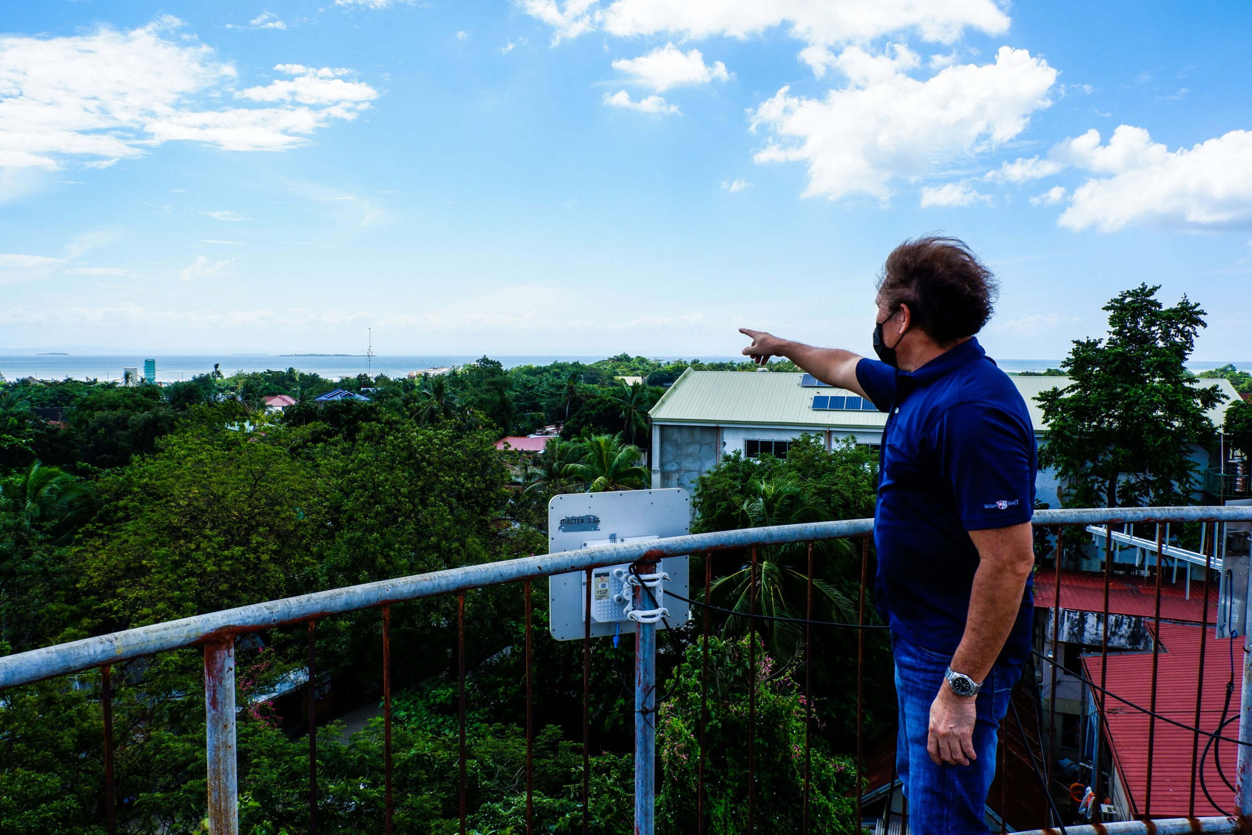 Jeff Llanto of the Central Visayas Information Sharing Network Foundation, Inc. (CVISNet) points to Gilutungan Island while on top of a water tank in the Cordova municipal hall. Attached to the railing beside him is a microwave dish that links the island to the town to synchronize data on a learning management system that runs independently in the remote barangay. That point-to-point connection also allows them to bridge the barangay to the high-speed internet they have in the town,